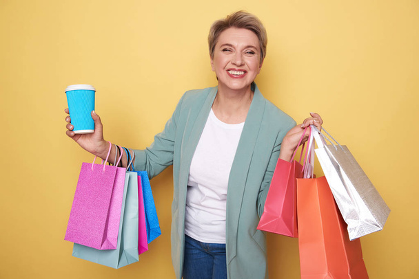 Happy woman looking at camera and holding colorful shopping bags and cupholder - 写真・画像