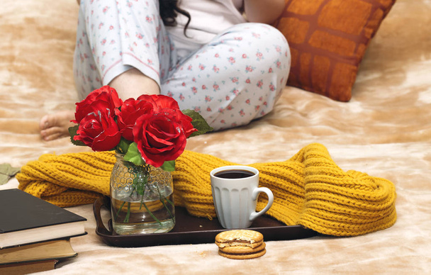 woman on the bed with old book and cup of coffee, roses vase, chocolate cookies in tray during the morning - Photo, Image