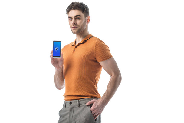 sonriente hombre con la mano en el bolsillo sosteniendo teléfono inteligente con aplicación shazam en la pantalla aislada en blanco
 - Foto, Imagen