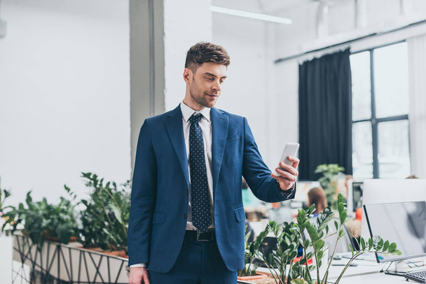 good-looking, smiling businessman in suite using smartphone in office - Valokuva, kuva