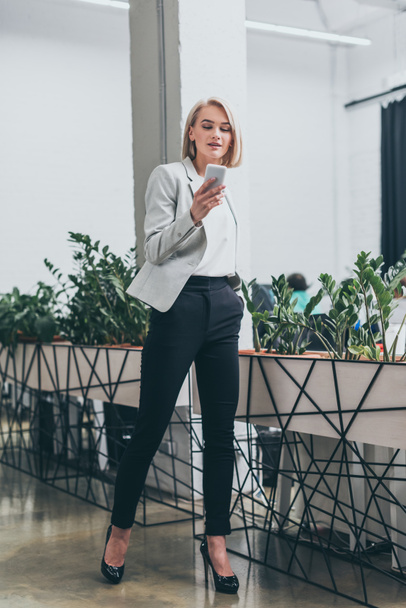 pretty businesswoman in formal wear and with hand in pocket using smartphone in office - Foto, Imagem