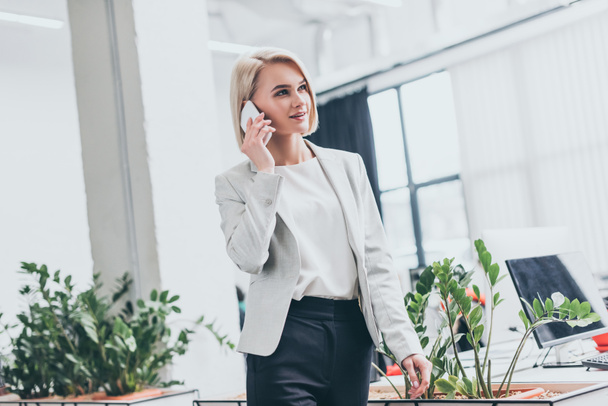 attractive blonde businesswoman in formal wear talking on smartphone in office - Фото, изображение