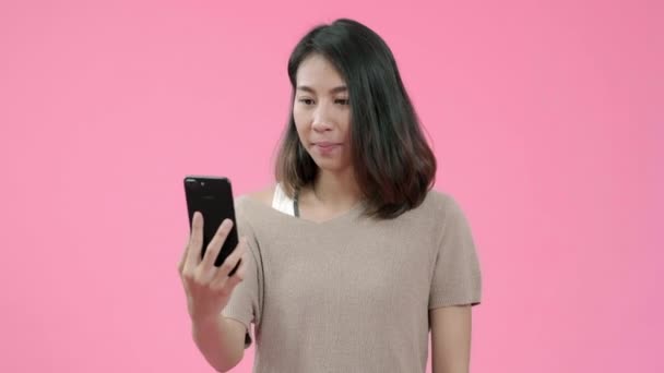 Young Asian woman using smartphone checking social media feeling happy smiling in casual clothing over pink background studio shot. Happy smiling adorable glad woman rejoices success. - Metraje, vídeo
