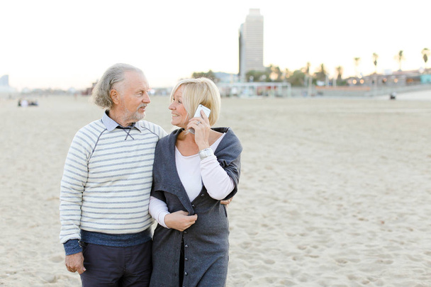 スマート フォンで話して金髪妻と砂浜の上を歩いてシニア夫. - 写真・画像