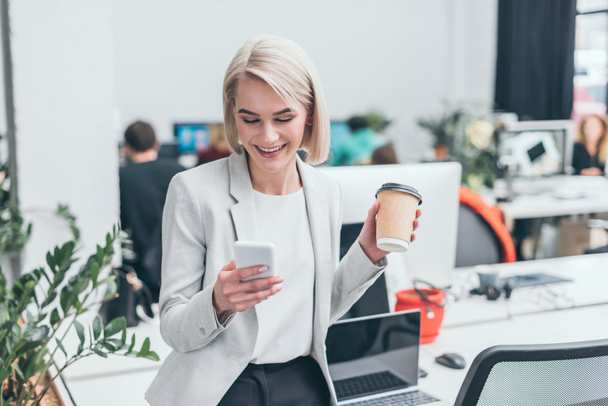 beautiful smiling businesswoman using smartphone and holding paper cup in office - Foto, Imagem