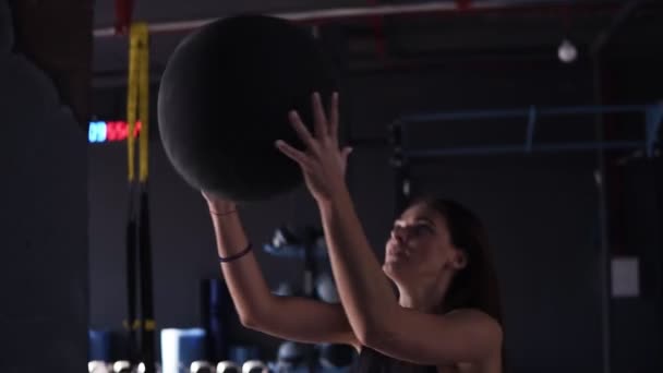 Active, young brunette woman doing squats while throwing a medicine ball up against a wall in a dark coloured modern gym full of equipment. Close up view - Filmmaterial, Video