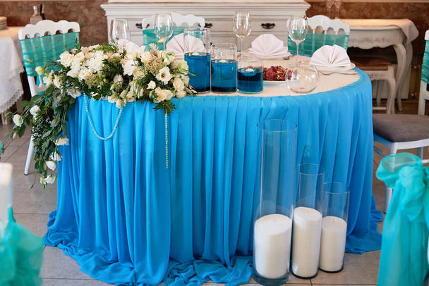 Wedding presidium in restaurant, free space. Banquet table for newlyweds with flowers, greenery, blue cloth and candles. Lush floral arrangement. Luxury wedding decorations - Photo, Image