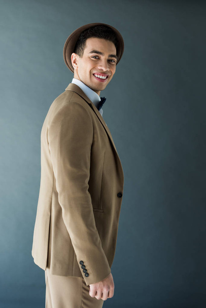 elegante hombre de raza mixta en traje mirando a la cámara y sonriendo aislado en gris
 - Foto, Imagen