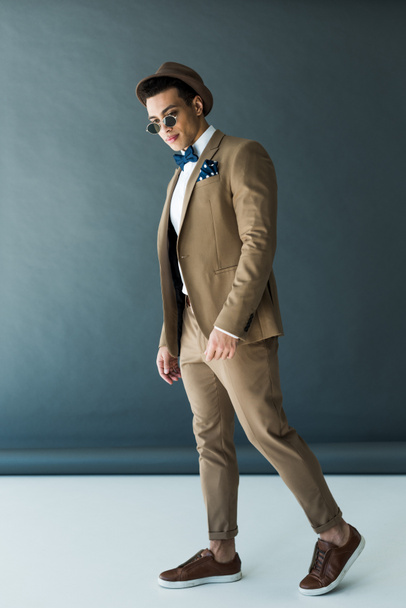 stylish mixed race man in suit, hat and sunglasses posing on grey - Foto, imagen