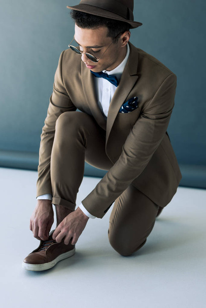 stylish mixed race man in suit and sunglasses putting on shoe on grey and white - Фото, изображение