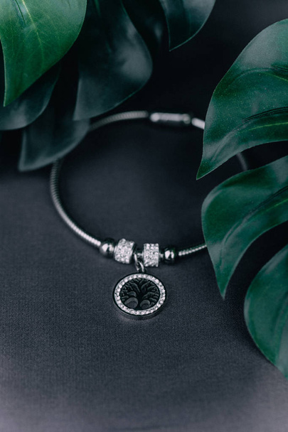 A close-up of a silver bracelet lies on a black background, next to flower sheets, on the bracelet there is a pendant in which is a tree. The concept of jewels. - Fotoğraf, Görsel