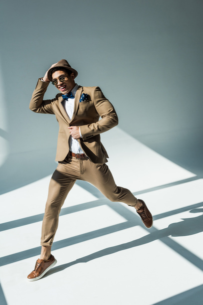 stylish mixed race man in suit smiling and jumping on grey with sunlight - Photo, Image