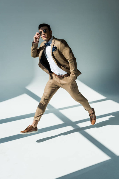 elegante hombre de raza mixta en traje y gafas de sol saltando sobre gris con luz solar
 - Foto, imagen