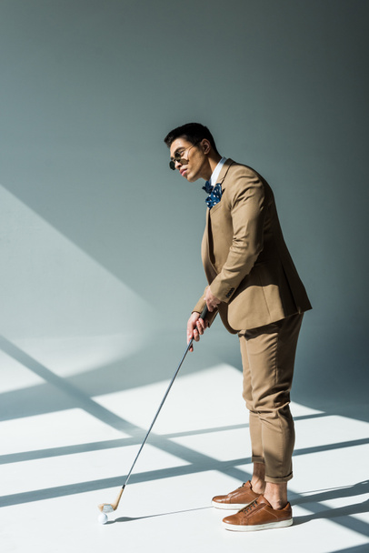 stylish mixed race man in suit holding golf club on grey with sunlight - Foto, imagen