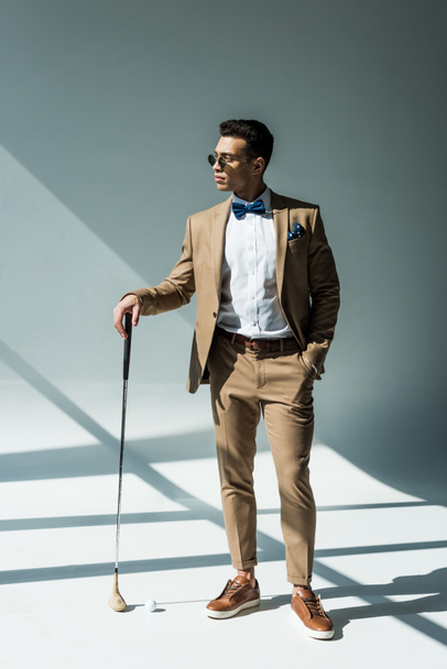 stylish mixed race man in suit posing with golf club on grey with sunlight - Foto, Imagen