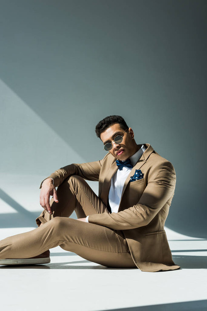 stylish mixed race man in suit and sunglasses sitting and posing on grey with sunlight and copy space - Foto, afbeelding