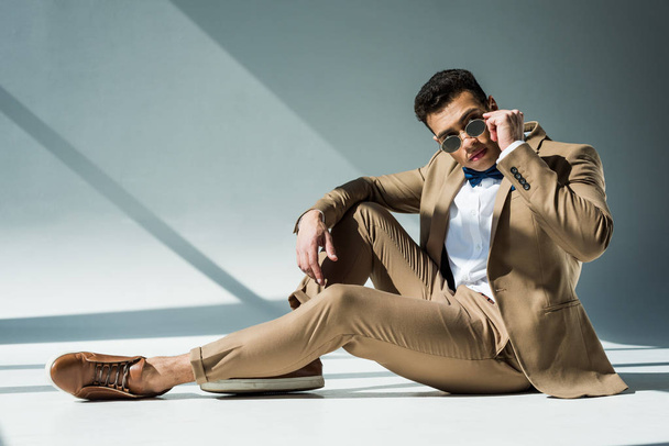 stylish mixed race man in suit and sunglasses sitting and posing on grey with sunlight and copy space - 写真・画像