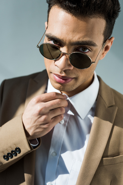 stylish mixed race man in suit and sunglasses adjusting shirt and looking at camera isolated on grey - Fotó, kép