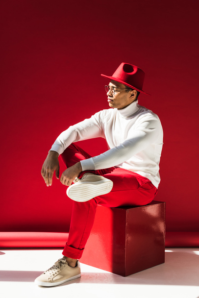 stylish mixed race man in hat and glasses sitting and posing on red - Photo, Image