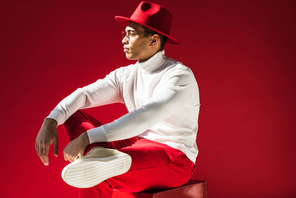 side view of stylish mixed race man in hat and glasses sitting and posing isolated on red - Foto, Imagem