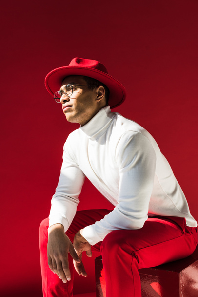 fashionable mixed race man in hat and glasses sitting and posing isolated on red - Foto, immagini
