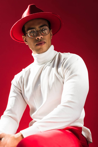fashionable mixed race man in hat and glasses sitting and posing isolated on red - Фото, изображение