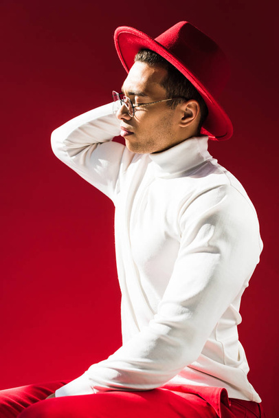stylish mixed race man in hat, turtleneck and glasses sitting and posing isolated on red - Foto, afbeelding
