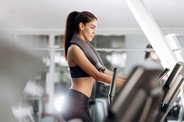 Dark-haired  athletic girl dressed in black sports top and tights with towel  on her neck works out on a treadmill in the gym - Foto, imagen