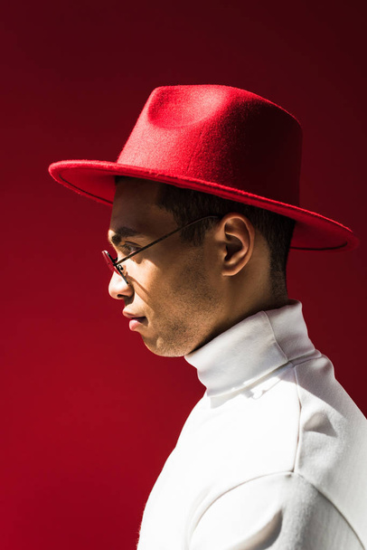 vista lateral do homem de raça mista elegante em chapéu e óculos posando isolado no vermelho
 - Foto, Imagem