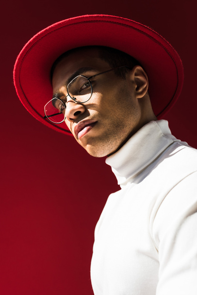 elegante hombre de raza mixta en sombrero y gafas mirando a la cámara mientras posando aislado en rojo
 - Foto, imagen