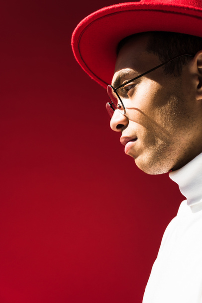 Zijaanzicht van stijlvolle mixed race man in Hat en glazen poseren geïsoleerd op rood met kopieerruimte - Foto, afbeelding