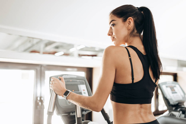 Dark-haired  athletic girl dressed in black sports top and tights works out on a treadmill in the gym - 写真・画像