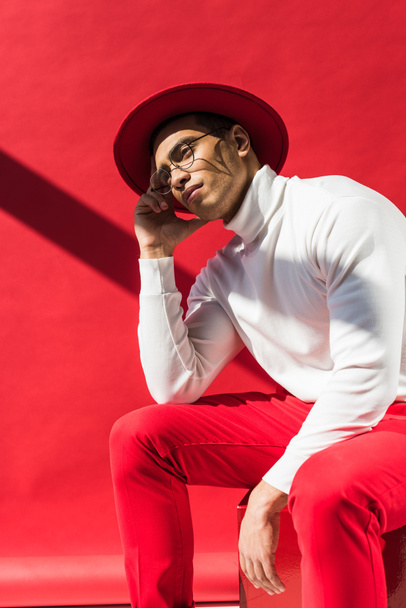 stylish thoughtful mixed race man in hat and glasses sitting and posing on red - Foto, Imagen
