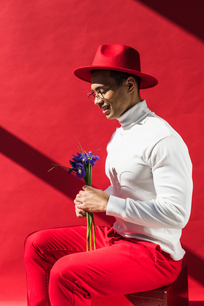 κομψό Μικτό άνθρωπο σε καπέλο και γυαλιά κρατώντας λουλούδια και χαμογελώντας στο κόκκινο - Φωτογραφία, εικόνα