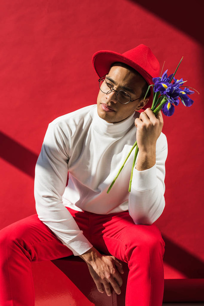 stylish mixed race man in hat and glasses holding flowers on red - Valokuva, kuva