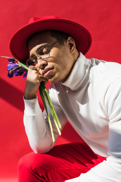 stylish mixed race man in hat and glasses holding flowers on red with shadow - Photo, image
