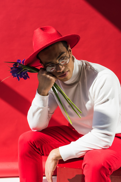 stylish mixed race man in hat and glasses holding flowers on red - Zdjęcie, obraz