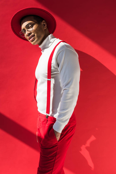 stylish mixed race man in hat and suspenders looking at camera while posing on red - Foto, Imagem