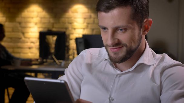 Nahaufnahme eines erwachsenen kaukasischen Geschäftsmannes, der auf dem Tablet SMS schreibt und am Arbeitsplatz im Büro fröhlich lächelt - Foto, Bild