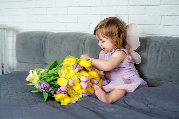 A menina com tulipas na roupa de fada senta-se em uma cama. Humor de primavera
 - Foto, Imagem