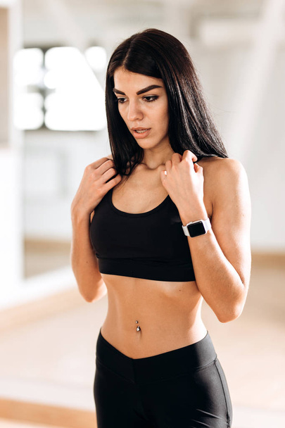 Beautiful slim girl dressed in black sportswear is standing in the gym in the fitness room next to the mirror - Fotoğraf, Görsel