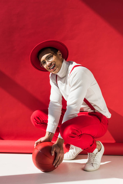 stylish mixed race man in hat and suspenders looking at camera while posing with basketball on red - Foto, afbeelding