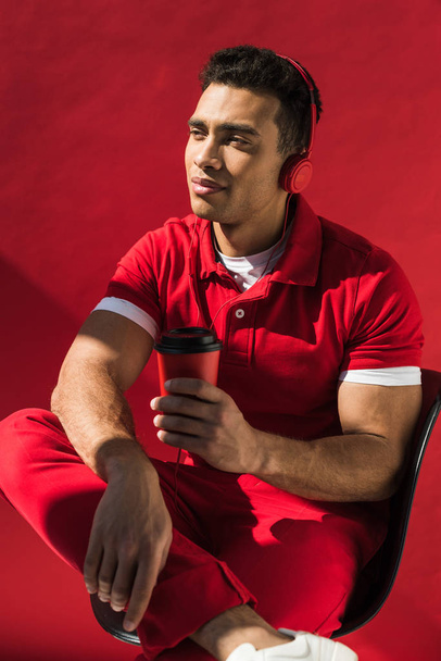 stylish mixed race man in headphones sitting, holding coffee to go and posing on red - Foto, immagini