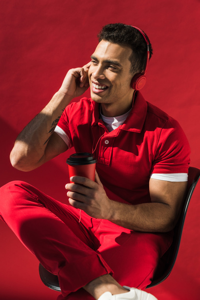 stylish mixed race man in headphones sitting, holding coffee to go and talking on smartphone on red - Foto, Imagem