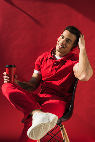 stylish mixed race man in headphones sitting, holding coffee to go and smiling on red - Zdjęcie, obraz