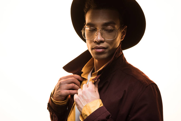 stylish mixed race man in glasses and hat adjusting coat and posing isolated on white - Foto, afbeelding