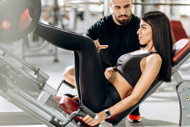 Athletic girl dressed in black sport clothes is lifting weights with her legs in the gym under the supervision of a coach - Foto, Imagem