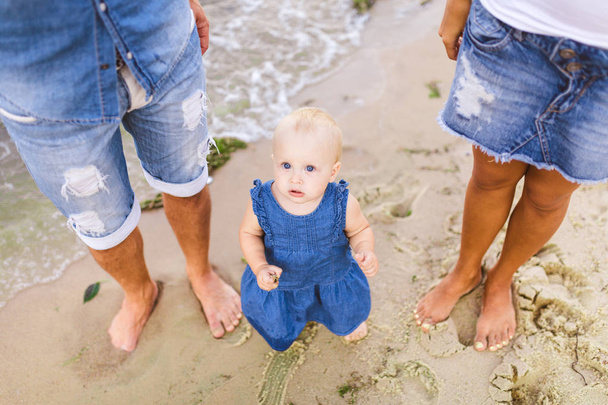 νεαρό ζευγάρι ετεροφυλόφιλων οικογένειας. Καυκάσιος μαμά και ο μπαμπάς μάθηση με τα πόδια ένα μικρό παιδί, κρατώντας τα χέρια ενός παιδιού σε μια αμμώδη παραλία κοντά στη θάλασσα. Οικογενειακές διακοπές στην παραλία, περπατώντας ξυπόλυτοι στη φύση - Φωτογραφία, εικόνα