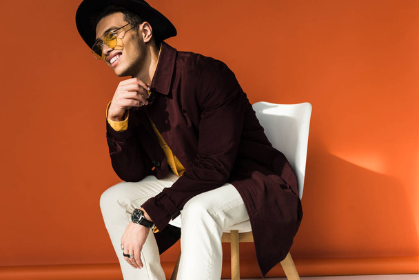 stylish mixed race man in hat and sunglasses sitting on chair and smiling on orange - Fotó, kép