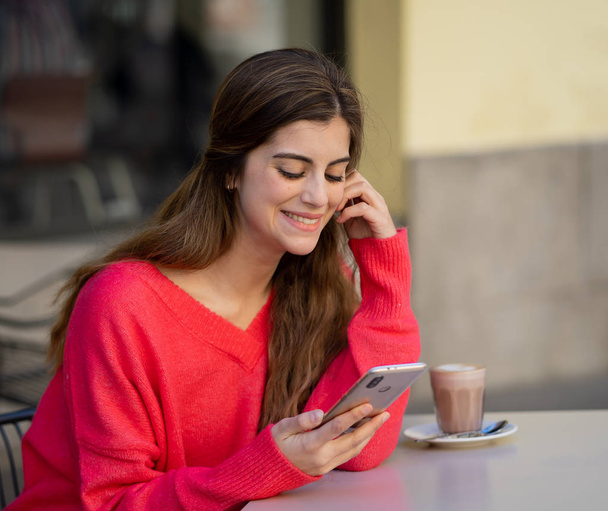 Mooie jonge vrouw met een rode trui, op zoek vrolijk een kopje koffie te drinken, chatten en praten over haar telefoon buiten op een koffieshop. Het gevoel van gelukkig en vrolijk. In levensstijl en technologie concept. - Foto, afbeelding